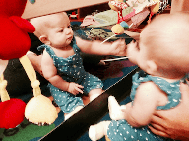 Baby in blue polka dot overalls looks in mirror.