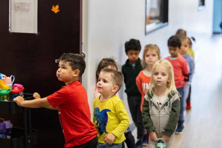 9 children are lined up and walking into their classroom. The child in the front of the line is pushing a cart filled with water bottles.